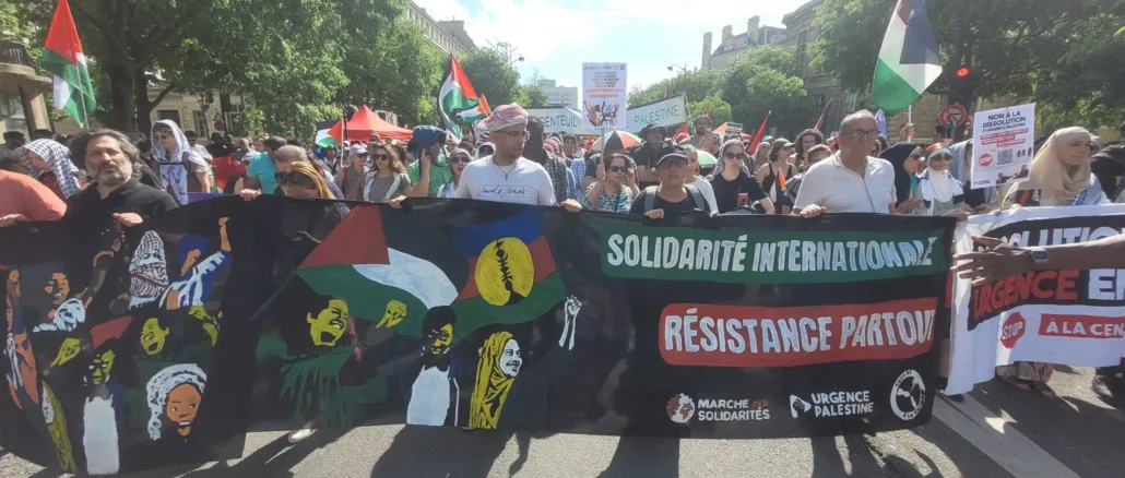 Sur une avenue ensoleillée et bordée d'arbres, un cortège de manifestant·es défile en tenant au premier plan une banderole qui représente 3 personnages en lutte avec des drapeaux palestiniens et kanak. la banderole est siglée "solidarité internationale, résistance partout", signée "marche des solidarités, urgence palestine et guerre à la guerre"