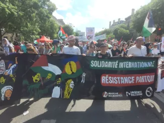 Sur une avenue ensoleillée et bordée d'arbres, un cortège de manifestant·es défile en tenant au premier plan une banderole qui représente 3 personnages en lutte avec des drapeaux palestiniens et kanak. la banderole est siglée "solidarité internationale, résistance partout", signée "marche des solidarités, urgence palestine et guerre à la guerre"
