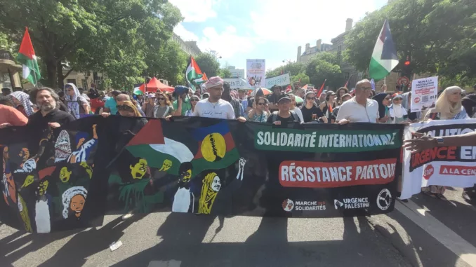 Sur une avenue ensoleillée et bordée d'arbres, un cortège de manifestant·es défile en tenant au premier plan une banderole qui représente 3 personnages en lutte avec des drapeaux palestiniens et kanak. la banderole est siglée "solidarité internationale, résistance partout", signée "marche des solidarités, urgence palestine et guerre à la guerre"