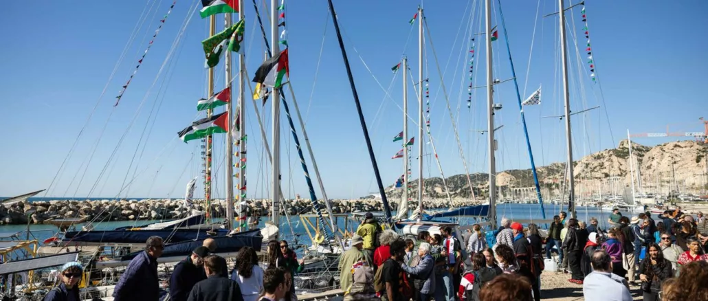 Sous un ciel bleu, une cinquantaine de personnes sont à quai devant une dizaine de voiliers dont les mâts et gréements sont ornés de drapeaux et fanions aux couleurs palestiniennes.