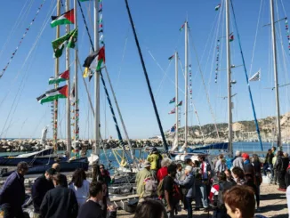 Sous un ciel bleu, une cinquantaine de personnes sont à quai devant une dizaine de voiliers dont les mâts et gréements sont ornés de drapeaux et fanions aux couleurs palestiniennes.