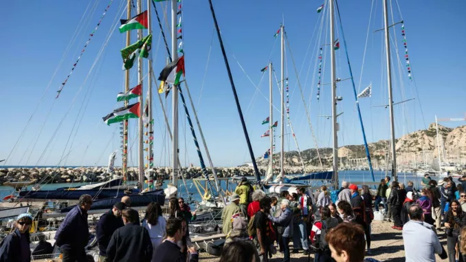 Sous un ciel bleu, une cinquantaine de personnes sont à quai devant une dizaine de voiliers dont les mâts et gréements sont ornés de drapeaux et fanions aux couleurs palestiniennes.